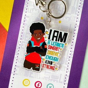 Peut inclure: Un porte-clés en acrylique représentant une illustration de dessin animé d'une jeune personne à la peau foncée et aux cheveux afro, portant un sweat à capuche rouge et une veste noire. Le texte indique : I AM A LEADER SMART BRAVE ENOUGH KIND STRONG.