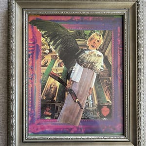 May include: Framed artwork featuring a collage of a person with a black wing and a sword, set against a backdrop of a building with columns. The artwork is framed in a silver-colored frame.