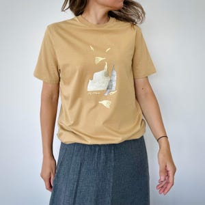 Könnte beinhalten: Ein beiges T-Shirt mit kurzen Ärmeln und einem zentralen Grafikdruck. Das Design besteht aus abstrakten Formen in Weiß- und Silbertönen, darunter eine texturierte, bergähnliche Form und verstreute blattartige Elemente auf der Brust.