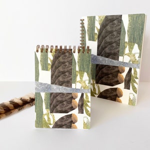 Può includere: Due quaderni con un design ispirato alla natura. Uno è rilegato a spirale, l'altro è con copertina rigida. Le copertine presentano un collage di forme verdi, marroni e grigie che ricordano alberi e fogliame. Una piuma è in primo piano.