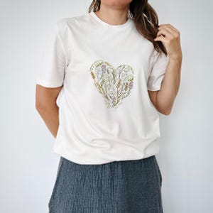 May include: A white short-sleeved t-shirt featuring a heart-shaped graphic composed of various botanical illustrations in muted green, yellow, and purple tones. The design is centered on the chest and includes small, delicate plant-like details.