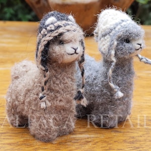 Puede incluir: Dos pequeñas figuritas de alpaca con gorros tejidos. La alpaca de la izquierda es marrón con un gorro blanco y negro. La alpaca de la derecha es gris con un gorro blanco.
