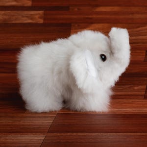 May include: A small, white plush elephant toy with a raised trunk and black eyes. The elephant is made of soft, fluffy material and is standing on a wooden surface.