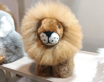 Op de afbeelding: Een pluche leeuwenknuffel met een bruin gezicht en een pluche manen. De leeuw zit op een lichtbruin oppervlak.
