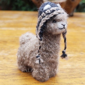 Puede incluir: Una pequeña figura de alpaca marrón que lleva un gorro de punto negro y blanco con largas borlas. La alpaca está de pie sobre una superficie de madera. Alpaca Retail.