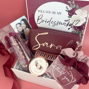 May include: A gift box containing a burgundy makeup bag with the name Sarah in gold, a glass with the name Sarah, a round compact mirror with a floral S, a hair claw, a bracelet with the text I CAN'T TIE THE KNOT WITHOUT YOU, and two scrunchies.