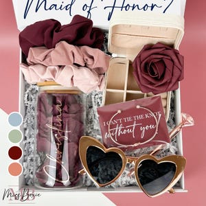 May include: A gift box filled with items for a maid of honor. The box includes a glass jar with a personalized message, a small jewelry box with a rose, heart-shaped sunglasses, hair scrunchies, and a card that says "I can't tie the knot without you!"