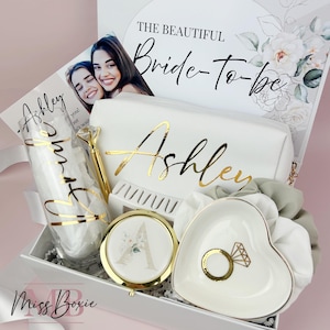 May include: A gift box containing a white pouch with gold text reading Ashley, a glass with gold text reading bride, a gold diamond-topped pen, a round compact mirror with a floral letter A, a heart-shaped trinket dish, and a white scrunchie.