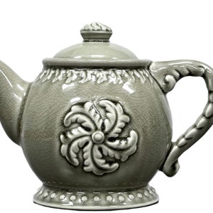 May include: A sage green ceramic teapot with a lid and handle. The teapot features a floral design on the front and a decorative handle. The teapot is approximately 7 inches tall and is suitable for serving tea.