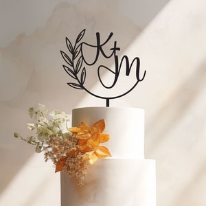 May include: A white two-tiered wedding cake with a black metal topper featuring a leafy wreath and the initials "K + M".