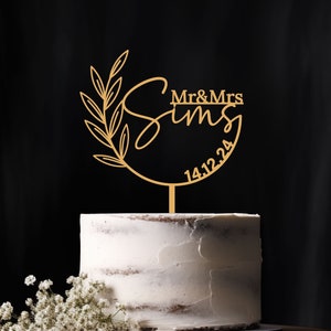 Peut inclure: Un topper de gâteau de mariage doré avec un motif de feuilles et le texte "Mr & Mrs Sims 14.12.24" sur un gâteau blanc.