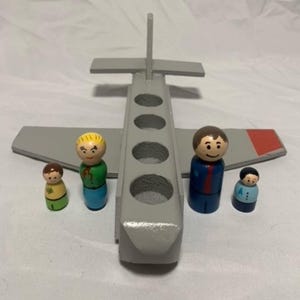 Pode incluir: Um avião de brinquedo de madeira cinza com quatro aberturas circulares, asas e uma barbatana caudal. Quatro pequenas bonecas de madeira em várias roupas estão perto do avião. A asa do avião tem um detalhe vermelho.