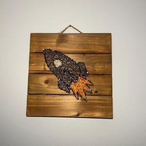 May include: A wooden wall hanging featuring a string art rocket ship design. The rocket is brown with a yellow and orange flame, mounted on a stained wooden plank background. A jute string is attached for hanging.