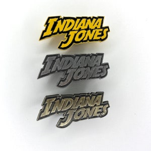 Puede incluir: Tres pines de metal con las palabras "Indiana Jones" en diferentes estilos. El pin superior es amarillo con letras negras, el pin del medio es plateado con letras negras y el pin inferior es dorado con letras negras.