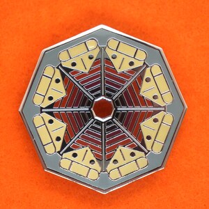Puede incluir: Un pin octogonal plateado con un diseño geométrico. El centro del pin es un círculo rojo rodeado de un patrón de rayas rojas y marrones alternas. El borde exterior del pin está decorado con ocho triángulos amarillos.