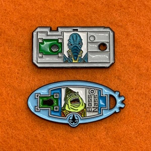 Könnte beinhalten: Zwei Emaille-Pins mit Figuren aus Star Wars. Der erste Pin ist silberfarben mit einem blauen Außerirdischen vor grünem Hintergrund. Der zweite Pin ist blau mit einem grünen Außerirdischen vor gelbem Hintergrund. Der Text "Boss Nass" befindet sich auf dem zweiten Pin.