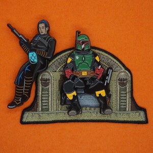 Könnte beinhalten: Eine grüne und schwarze Boba Fett Actionfigur sitzt auf einem Thron mit einer blauen und schwarzen Figur, die hinter ihm steht. Der Thron ist aus braunem Stoff mit einem grün-braunen Muster.