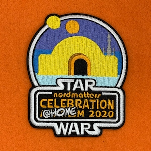 May include: A Star Wars Celebration patch with a yellow and blue image of a building with a black door, a yellow sun, and a blue sky. The text "STAR WARS" is at the bottom of the patch, and the text "nerdmatters CELEBRATION @HOME 2020" is in the middle of the patch.