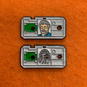 May include: Two silver-colored metal pins with a white background. The first pin features a cartoon image of George Lucas, the creator of Star Wars. The second pin features a cartoon image of Chewbacca, a Wookiee character from Star Wars.