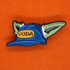 May include: A blue and green enamel pin featuring a baseball cap with the word "YODA" on the front. The cap has a green, pointed ear sticking out from the side.