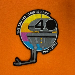 May include: A circular embroidered patch featuring a gray and black AT-AT walker from the movie "The Empire Strikes Back" with the text "The Empire Strikes Back 40 1980-2020".