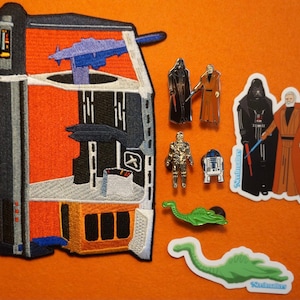 May include: A set of Star Wars themed embroidered patches and pins. The patches feature Darth Vader, Obi-Wan Kenobi, and a spaceship. The pins feature a Stormtrooper, R2-D2, and a character from the Star Wars universe.
