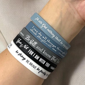 Può includere: Una pila di sei braccialetti in silicone in varie tonalità di blu, grigio e bianco. Ogni braccialetto presenta una citazione ispiratrice in lettere bianche. I braccialetti sono impilati su un polso, con il braccialetto superiore che recita "For with God nothing shall be impossible."
