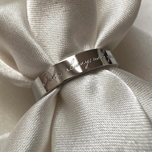 May include: A silver ring with the engraved text "God is always with you." The ring is displayed on a white, silky fabric. The ring has a flat, polished surface and a simple, elegant design.