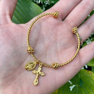 May include: A gold-toned twisted bangle bracelet with four knotted accents and two charms: a cross and a depiction of the Virgin Mary.