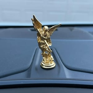 May include: A gold-colored hood ornament depicting a winged figure with outstretched wings. The figurine is mounted on a dark surface, likely the hood of a vehicle. The ornament has intricate details and a polished finish.