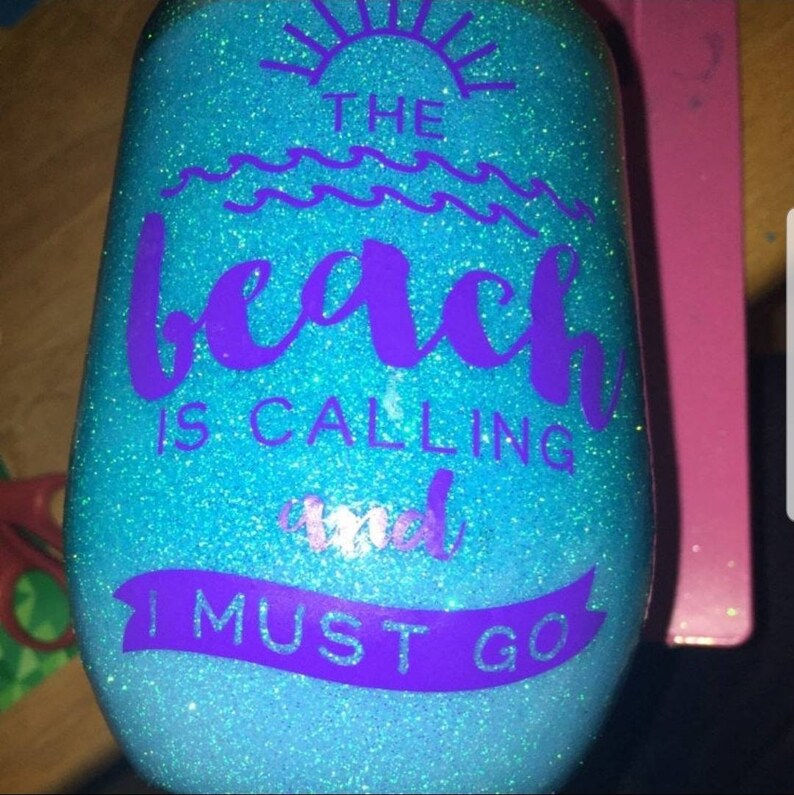 May include: A stemless wine glass with a glittery blue and turquoise finish. The text "THE beach IS CALLING and I MUST GO" is in purple. The design includes a sun and waves.