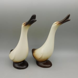 May include: Two ceramic duck figurines with brown and white glaze. The ducks have their heads tilted back and their beaks open.