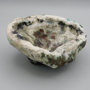 May include: A small, handmade, irregularly shaped bowl with a rough, textured surface. The bowl is primarily off-white with areas of black, green, and reddish-brown coloration, suggesting an aged or weathered appearance. The interior is a muted mix of colors.
