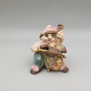 May include: A ceramic figurine of a person wearing a pink hat and blue pants playing a brown and yellow cello.