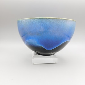 May include: A ceramic bowl with a glossy, layered glaze in shades of blue and black. The bowl has a rounded shape with a slightly flared rim, and is displayed on a white stand. The rim has a thin, light green band.