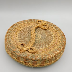 May include: A round, handwoven basket with a lid. The basket is made of natural-colored fibers, with a braided handle on the lid. The weave pattern is intricate, with a spiral design on the lid's center. The basket is likely used for storage or decoration.