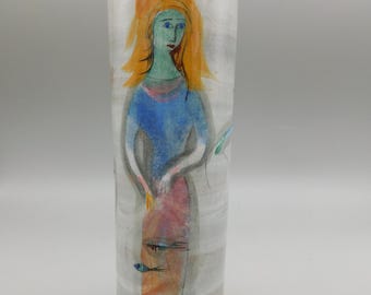 May include: A tall, cylindrical ceramic vase with a hand-painted illustration of a woman. The woman has orange hair, a blue top, and a pink skirt. Two small fish are painted near the bottom. The vase is approximately 10 inches tall.