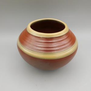 May include: A round, ceramic vase with a wide opening. The vase is primarily reddish-brown with a horizontal band of light yellow and a darker brown interior. The exterior has a textured, ribbed design.