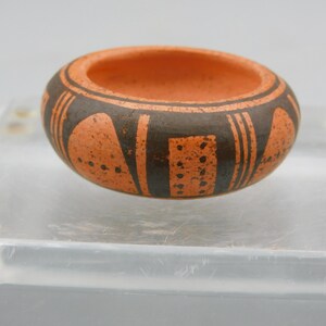May include: A small, round, terracotta-colored ceramic bowl featuring a matte finish. The exterior is decorated with dark brown geometric patterns, including vertical lines, solid shapes, and small dots arranged in a repeating design around the rim.