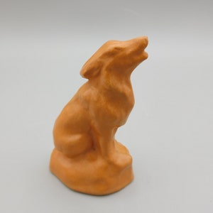 May include: A small, orange ceramic figurine of a wolf howling at the moon. The wolf is sitting on a small base.