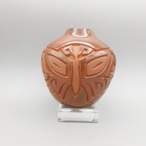 May include: A terracotta-colored ceramic vase featuring a carved butterfly motif on its rounded body. The vessel has a smooth, polished finish and a narrow opening at the top. It is displayed resting on a clear, rectangular acrylic base.