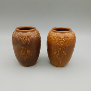 May include: Two matching brown ceramic vases with a rounded body and a narrow opening. Each vase features a raised, embossed floral pattern around the upper section, consisting of stylized petals and small circular details in a monochromatic finish.