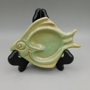 May include: A ceramic fish-shaped dish in shades of green and yellow, resting on a black stand. The dish features a detailed eye and fin design, perfect for holding small items. A decorative accent for any home.