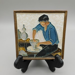 May include: A ceramic tile with a painted image of a person making pottery on a wheel. The person is wearing a blue shirt and black pants. The tile has a brown frame.