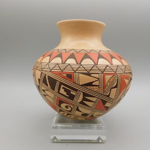 May include: A handcrafted, round pottery vase with a beige body and a flared rim. The vase features intricate geometric patterns in black and red-orange. The design includes triangles, lines, and shapes. The vase is displayed on a clear acrylic stand.