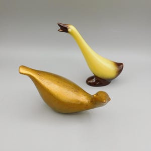 May include: Two ceramic bird figurines. One is gold-colored, the other is yellow with brown accents. The yellow bird has an open beak. Both have a smooth, glossy finish. Decorative home decor.