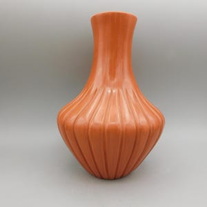 May include: An orange ceramic vase with a ribbed design. The vase has a bulbous base that tapers to a narrow neck and flared opening. The vase is a solid color and has a glossy finish. The vase is a decorative item.