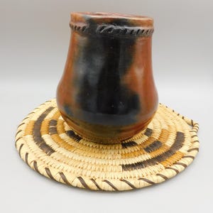 May include: A handmade ceramic vase with a dark brown and reddish-brown color scheme. The vase sits on a round, woven straw mat with brown and tan stripes. The vase has a textured rim.