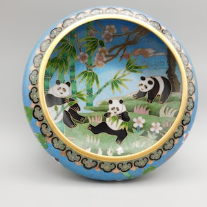 May include: A round, decorative dish with a blue exterior and a gold rim. The interior features a scene with three pandas, bamboo, and flowering trees. The pandas are black and white, and the background is a light blue.