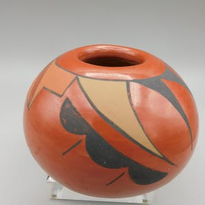May include: A round, terracotta-colored ceramic vase with a small opening at the top. The vase is adorned with geometric designs in black, tan, and orange. The design includes curved and angular shapes. The vase is displayed on a clear stand.